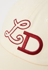 Kremfarget caps med mørkerøde broderte bokstaver "L" og "D" med dekorative svulster, laget av teksturert stoff, detaljert søm.