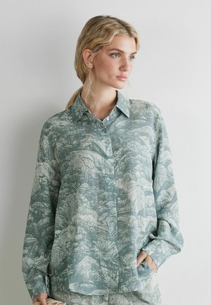 Vrouw met blond haar in een lage paardenstaart, gekleed in een losvallend overhemd met lange mouwen en een lichtgroen tropisch bladerenpatroon.