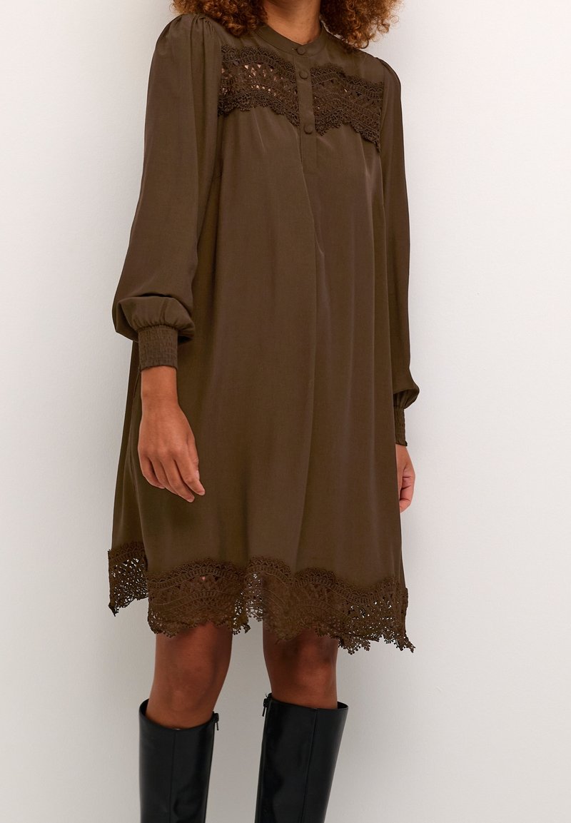 Femme portant une robe marron ample avec des détails en dentelle sur la poitrine et l'ourlet, des manches longues bouffantes et des bottes noires montantes jusqu'aux genoux.