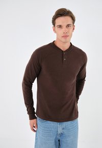 Cămașă henley maro cu mânecă lungă, cu butoni și manșete cu model în relief, combinată cu pantaloni scurți din denim de culoare bleu deschis. Textură fină și croială lejeră.