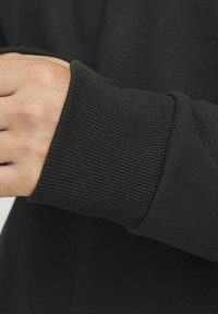 Primo piano di un tessuto di felpa nera, con polsino a coste e superficie liscia. La manica è parzialmente arrotolata per rivelare il braccio.
