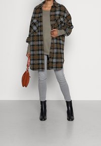 Person, die eine übergroße karierte Jacke, graue Jeans, schwarze Stiefeletten, ein olivfarbenes Langarmshirt trägt und eine braune Handtasche vor einem schlichten Hintergrund hält.