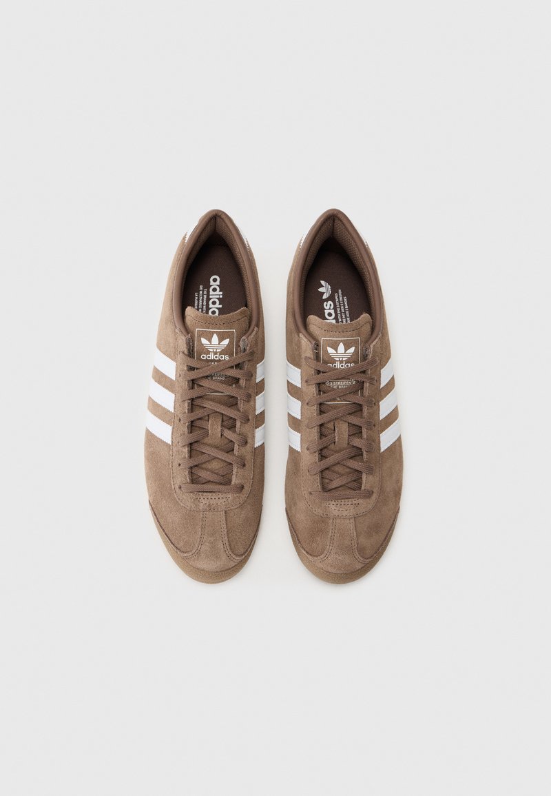 Baskets en daim marron avec un design à trois bandes blanches et un logo. Présentent une languette rembourrée et des lacets, offrant un look athlétique classique.