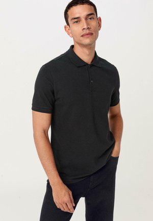 Schwarzes Poloshirt aus weichem Material mit einer Knopfleiste mit drei Knöpfen, kurzen Ärmeln und einer leicht taillierten Silhouette. Kombiniert mit dunklen Jeans.