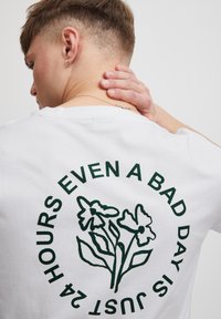 Hvid bomulds t-shirt med et grønt trykt blomstermotiv og teksten "SELV EN DÅRLIG DAG ER BARE 24 TIMER" i en cirkulær opstilling.