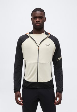 Bărbat purtând un hanorac cu fermoar negru și bej cu logo pe piept și mânecă, stând cu brațele relaxate pe un fundal simplu.