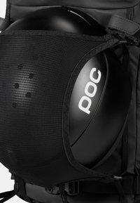 En svart hjelm som er sikret i en nettinglomme, med en blank finish og en hvit "POC" logo. Bakgrunnen er et strukturert svart ryggsekkstoff.