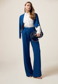 Abito blu su misura con blazer aderente, pantaloni a gamba larga e camicia bianca. Accentuato da una clutch con pietre preziose e orecchini audaci. Texture liscia.