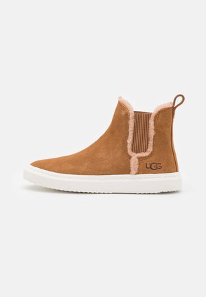 Bruine suède instap enkellaars met witte zool, bontvoering rand, elastische zijpanelen en UGG-logo op de hiel.