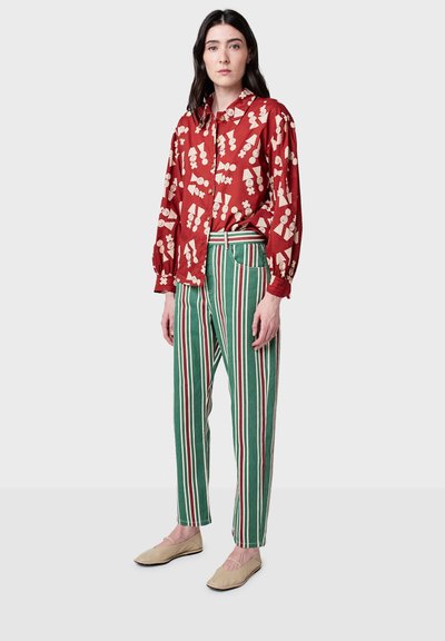 Camicia rossa a fantasia con maniche a palloncino, abbinata a pantaloni a righe verdi e bianche. Scarpe ballerine beige completano il look.