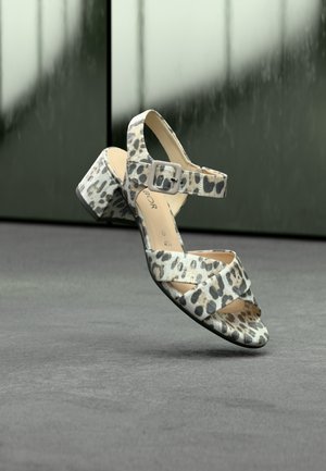 Slipper met luipaardprint, blokhak en enkelriem, zwevend boven een grijze oppervlakte met een wazige donkere achtergrond.