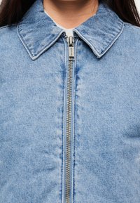 Ανοιχτό μπλε τζιν μπουφάν με γιακά και κεντρικό φερμουάρ, με υφή επιφάνειας και ορατές ραφές.