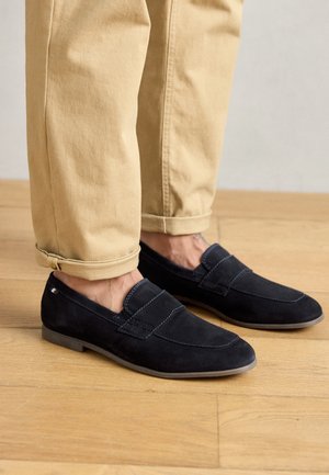 Fødder iført sorte ruskind loafers kombineret med op rullede beige bukser på et lyst trægulv.