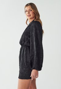 Schwarzes Kleid aus gewelltem Stoff, mit tiefem Ausschnitt, langen Puffärmeln und taillierter Taille. Glatte Textur und kurze Länge.