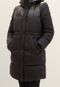 Długi czarny pikowany płaszcz zimowy z zatrzaskami, kieszeniami z klapami i wysokim kołnierzem, noszony przez osobę z rękami w kieszeniach.