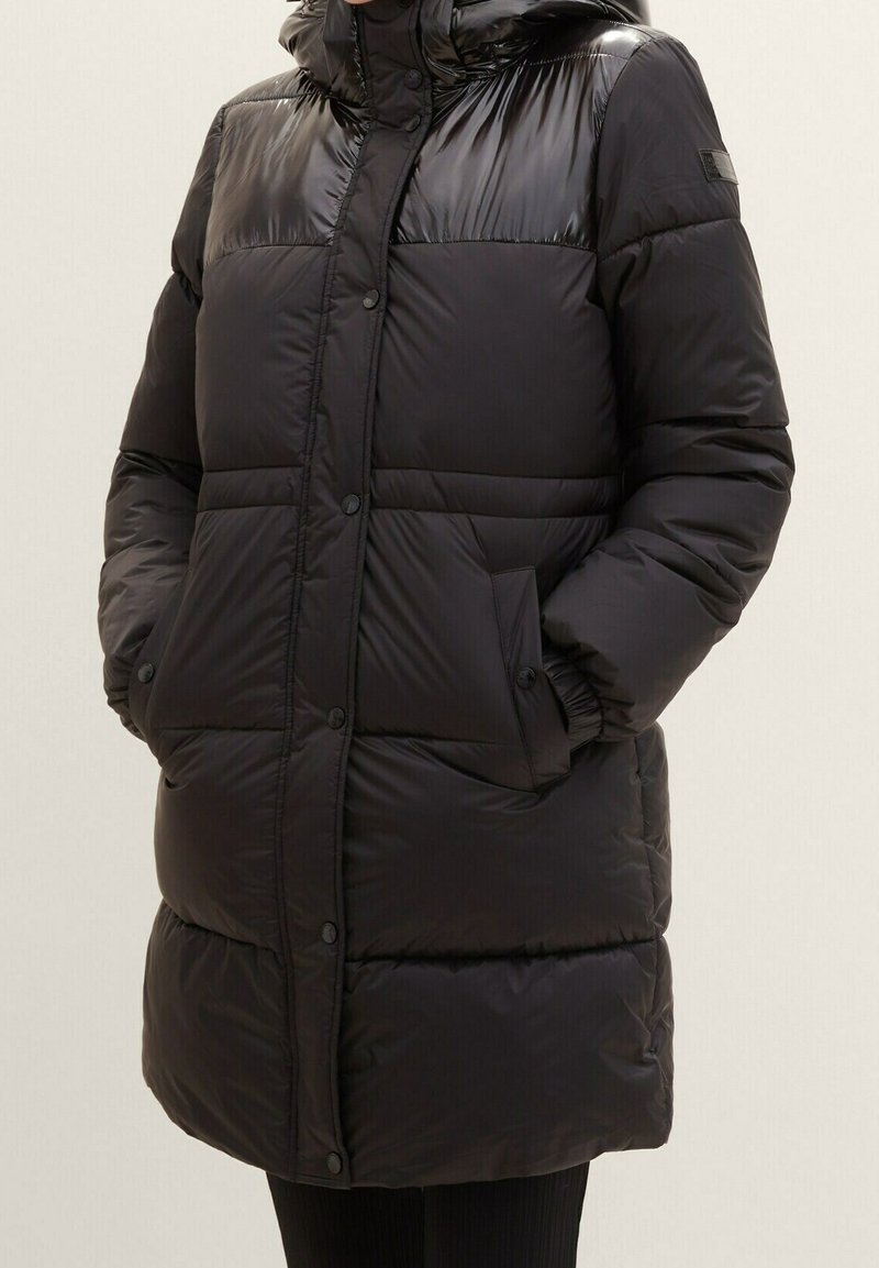 Długi czarny pikowany płaszcz zimowy z zatrzaskami, kieszeniami z klapami i wysokim kołnierzem, noszony przez osobę z rękami w kieszeniach.