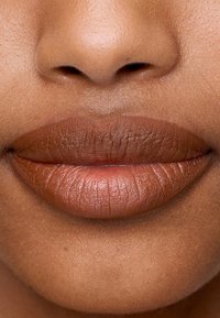 Nærbilde av fyldige lepper med matt finish i en varm, dempet brunfarge. Teksturert overflate med subtile linjer og naturlige konturer.