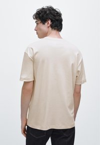 Beige t-shirt med korta ärmar, rund halsringning och ledig passform, visad bakifrån mot en enfärgad bakgrund. Mjuk tygstruktur.