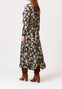 Černé květinové šaty s žlutými a bílými květy, dlouhými rukávy, staženým páskem a volánkovým lemem, doplněné hnědými semišovými kozačkami po kolena.