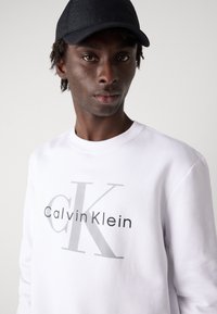 Felpa bianca in cotone con un grande logo grigio "Calvin Klein" e dettagli neri, indossata con un cappellino nero. Tessuto morbido, vestibilità comoda.
