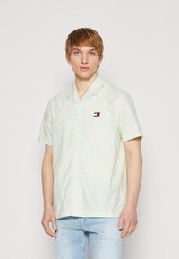 Camisa de manga corta, de color verde claro, con patrones abstractos amarillos, con botones en la parte delantera y un pequeño logo en el pecho. Fabricada con una tela ligera.