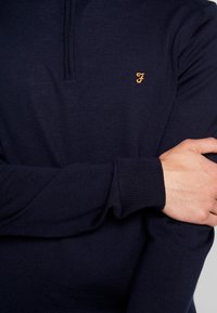 Mörkblå stickad tröja med halvdragkedja, ribbade ärmslut och en broderad logotyp i orange på bröstet. Mjuk och texturerad material.