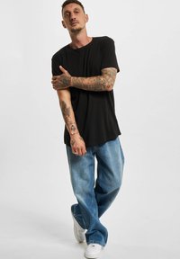 Svart kortermet t-skjorte kombinert med lyseblå løstsittende denimjeans og hvite joggesko. Synlige tatoveringer på armene. Enkel bakgrunn.