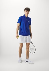 Blå polo skjorte med krage og logo, kombinert med hvite shorts. Modellen holder en tennisball og racket, iført hvite joggesko.