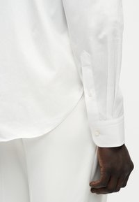 Χέρι με σκούρο δέρμα που ακουμπάει κοντά στο ισχίο, φορώντας ένα λευκό πουκάμισο με μακριά μανίκια με μανσέτα με κουμπιά και λευκό παντελόνι σε μονόχρωμο φόντο.