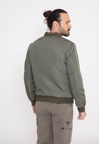Olijfgroene bomberjack met geribbelde zoom en cuffs, met een gladde, glanzende textuur en minimale stikseldetails.