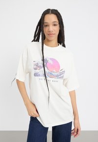T-shirt blanc surdimensionné avec un graphisme "La Grande Vague" présentant des vagues bleues et un soleil rose. Manches courtes, matériau en coton, coupe décontractée.
