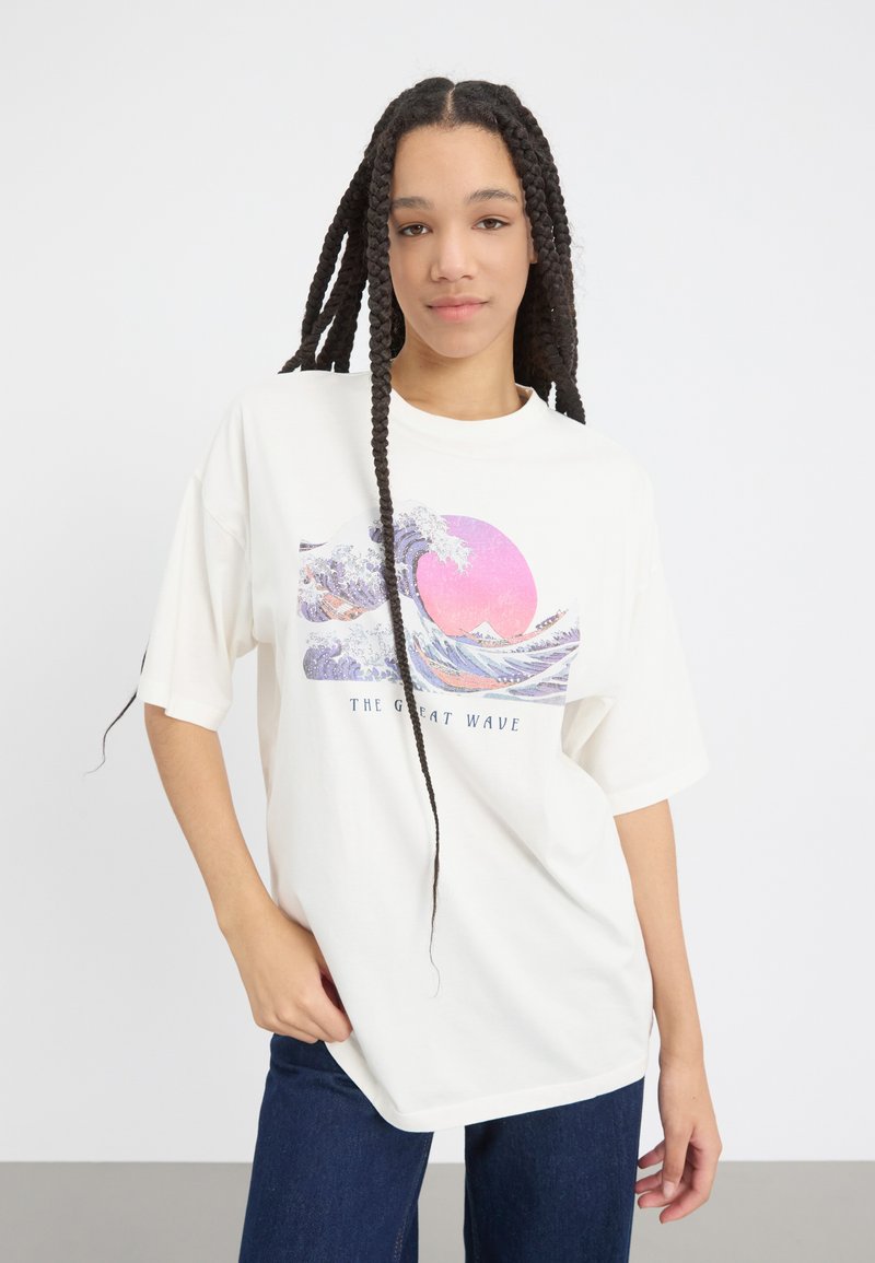 T-shirt blanc surdimensionné avec un graphisme "La Grande Vague" présentant des vagues bleues et un soleil rose. Manches courtes, matériau en coton, coupe décontractée.