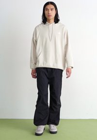 Sweat-shirt beige à capuche avec une poche frontale, associé à un pantalon noir ample. Le modèle porte des baskets gris clair. L'arrière-plan est blanc uni.