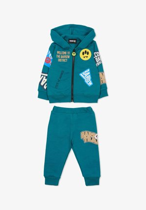 Teal twee-delige hoodie en sweatpants set gemaakt van zacht materiaal. De hoodie heeft graphics en een rits; de broek heeft een opdruk met logo op het been.