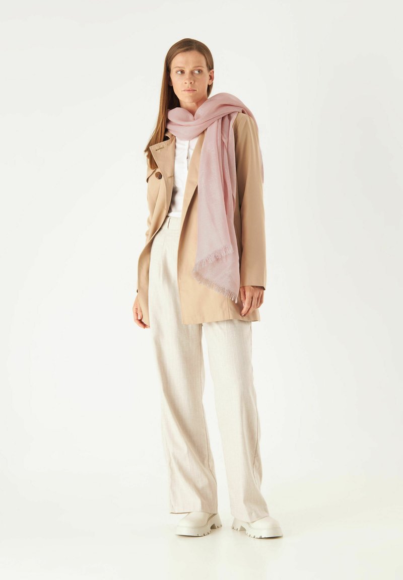 Manteau beige, haut blanc, pantalon gris clair et écharpe rose à bords frangés. Des chaussures blanches imposantes complètent la tenue. Design minimaliste.