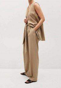 Kobieta w luźnej beżowej bezrękawnik i pasujących szerokich spodniach, w brązowych sandałach, stojąca z jedną ręką w kieszeni.