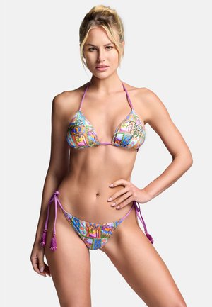 Vrouw poseert in een kleurrijke, patroon bikini met paarse tiestraps tegen een effen witte achtergrond.