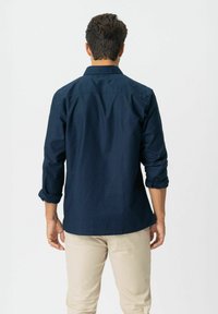 Man die met zijn rug naar de camera staat, gekleed in een marineblauwe langemouwen shirt met opgerolde mouwen en beige broek tegen een effen achtergrond.