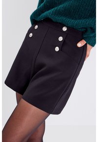 Pantalones cortos negros hechos de una tela suave, con botones decorativos plateados, bolsillos laterales y un corte ajustado. Llevados con medias negras.