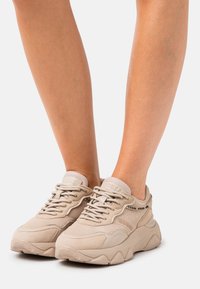 Beige sneakers featuring a textured mesh upper, suede accents, thick sole, and tonal laces. Brand logo visible on the side.