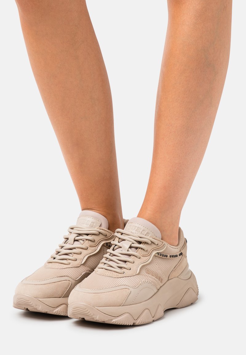 Beige sneakers featuring a textured mesh upper, suede accents, thick sole, and tonal laces. Brand logo visible on the side.