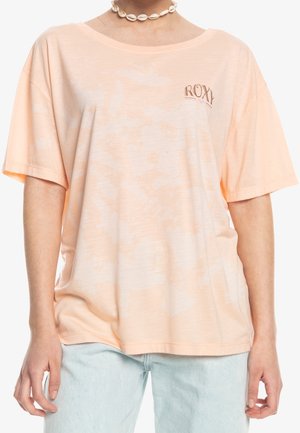 Femme portant un t-shirt ample tie-dye pêche avec le logo « ROXY » sur la poitrine gauche, un jean bleu clair et un collier de coquillages blanc.