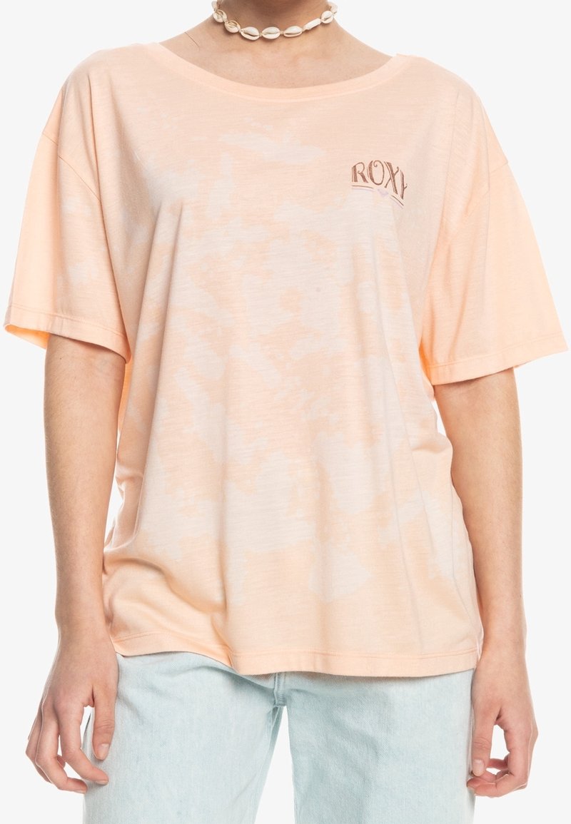 Femme portant un t-shirt ample tie-dye pêche avec le logo « ROXY » sur la poitrine gauche, un jean bleu clair et un collier de coquillages blanc.