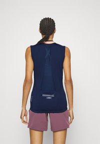 Personne ayant des cheveux tressés portant un gilet de sport sans manches bleu marine avec une poche à fermeture éclair et un short de sport mauve, vue de derrière.