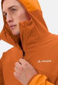 Veste légère orange avec un col haut, fermeture éclair à l'avant et logo. Présente une texture lisse et des poignets élastiques.
