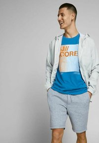 Giacca zip grey chiaro sopra una t-shirt grafica blu con testo arancione, abbinata a pantaloni corti grigi. Il tessuto sembra morbido, look sportivo e casual.