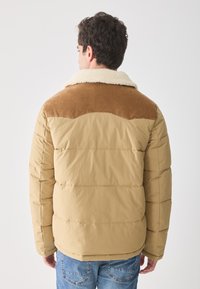 Hombre visto de espaldas que lleva una chaqueta acolchada beige con un yugo de pana marrón y un cuello de borreguito, combinada con vaqueros azules.