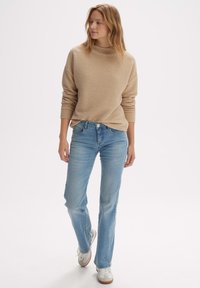 Beige gebreide trui met een hoge nek, gecombineerd met lichtblauwe recht-toe-rechtaan jeans en witte sneakers met bruine accenten.