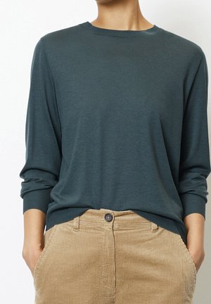 Personne portant une chemise à manches longues vert foncé rentrée dans un pantalon beige en velours côtelé, les mains dans les poches, cadrée au niveau du cou et des hanches.