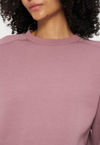 Femme aux cheveux bouclés et foncés portant un pull mauve à col rond, montrée des épaules jusqu’au milieu du visage sur un fond blanc.